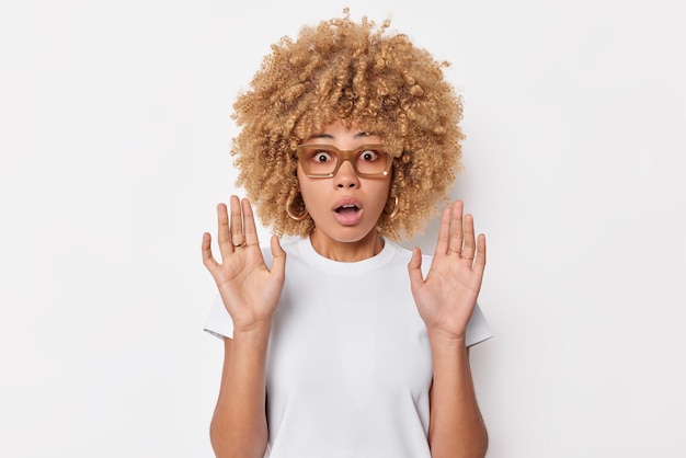 Schockierte, sprachlose, lockige Frau hält die Handflächen in defensiver Pose erhoben, sieht etwas Erschreckendes, hält den Atem vor Staunen an, trägt eine Brille und ein lässiges T-Shirt, das über weißem Hintergrund isoliert ist