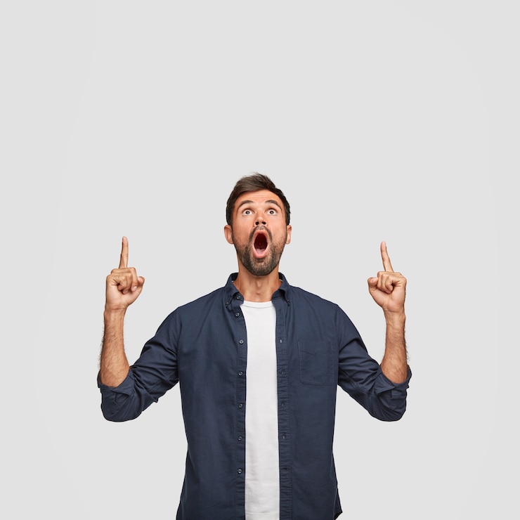 Kostenlose Foto schockierter bärtiger kerl mit überraschtem blick, zeigt mit beiden zeigefingern oben, wundert sich, als er etwas unglaubliches sieht