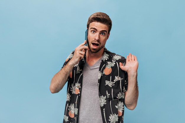 Schockierter bärtiger Typ mit blauen Augen im modernen Sommerhemd spricht am Telefon und schaut überrascht in die Kamera vor isoliertem Hintergrund