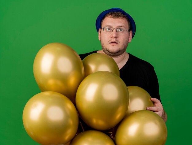 Schockierter erwachsener slawischer Mann in optischer Brille mit blauem Partyhut hält Heliumballons nach oben