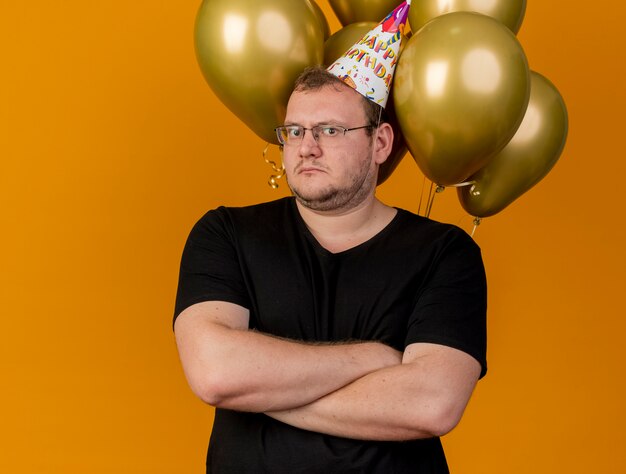 Schockierter erwachsener slawischer Mann in optischer Brille mit Geburtstagsmütze steht mit verschränkten Armen vor Heliumballons
