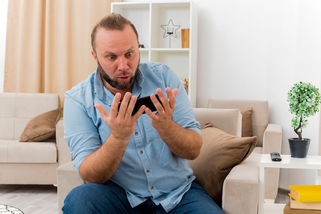 Schockierter erwachsener slawischer Mann sitzt auf Sessel, der Telefon im Wohnzimmer hält und betrachtet