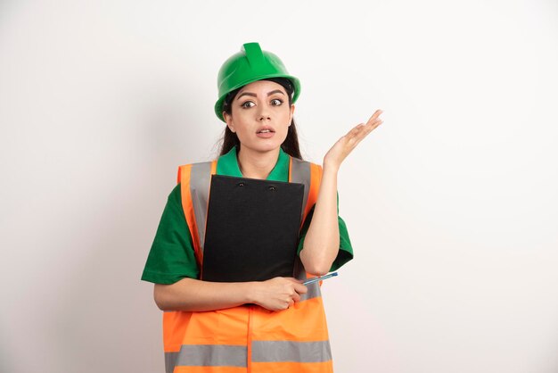 Schockierter frauenkonstrukteur mit grünem helm, der mit zwischenablage steht. foto in hoher qualität