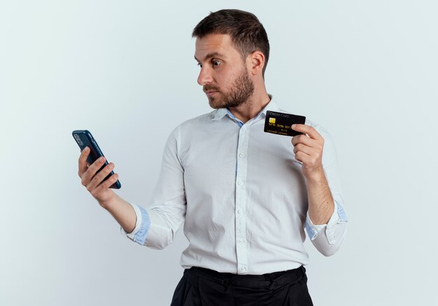 Schockierter gutaussehender Mann hält Kreditkarte, die Telefon lokalisiert auf weißer Wand betrachtet