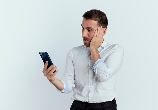 Schockierter gutaussehender Mann legt Hand auf Gesicht, das Telefon lokalisiert auf weißer Wand betrachtet