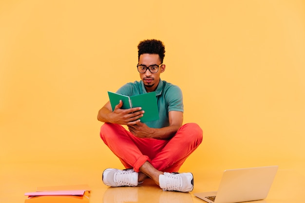 Schockierter internationaler student, der mit lehrbuch auf dem boden sitzt. innenaufnahme des beschäftigten männlichen freiberuflers, der nahe laptop aufwirft.