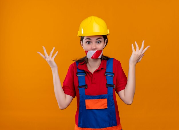 Schockierter junger Baumeistermädchenmund, der mit Warnband versiegelt wird, hebt Hände auf lokalisiertem orangefarbenem Hintergrund an