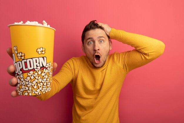 Schockierter junger blonder gutaussehender Mann, der einen Eimer Popcorn ausstreckt und die Hand auf den Kopf legt