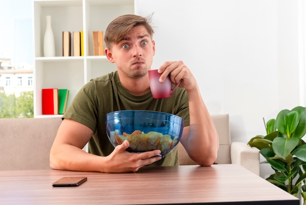 Schockierter junger blonder gutaussehender Mann sitzt am Tisch mit Telefon, das Schüssel Chips und Tasse innerhalb des Wohnzimmers hält