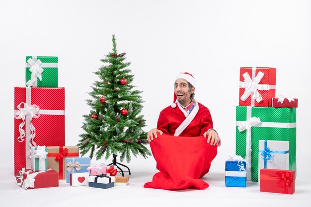 Schockierter junger Erwachsener verkleidet als Weihnachtsmann mit Geschenken und geschmücktem Weihnachtsbaum, der auf dem Boden auf weißem Hintergrund sitzt