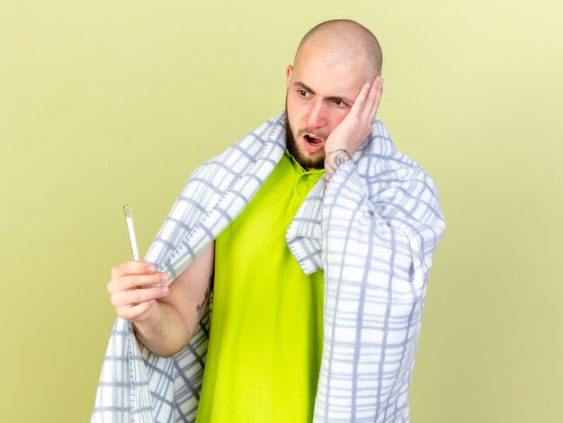 Schockierter junger kranker Mann, der in Plaid gewickelt ist, legt Hand auf Gesicht, das Thermometer hält und auf olivgrüne Wand isoliert betrachtet