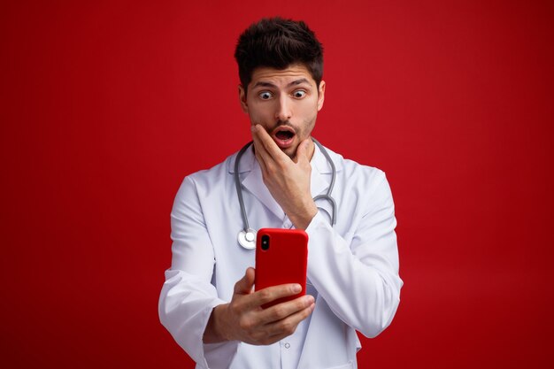 Schockierter junger männlicher Arzt, der eine medizinische Uniform und ein Stethoskop um den Hals trägt und ein Handy hält, das die Hand am Kinn hält und per Videoanruf isoliert auf rotem Hintergrund spricht