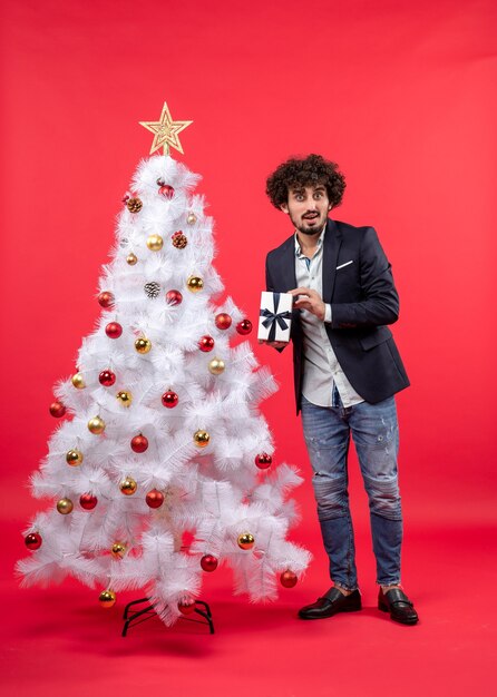 Schockierter junger Mann, der sein Geschenk hält, das nahe geschmücktem weißem Weihnachtsbaum auf der rechten Seite von Rot steht