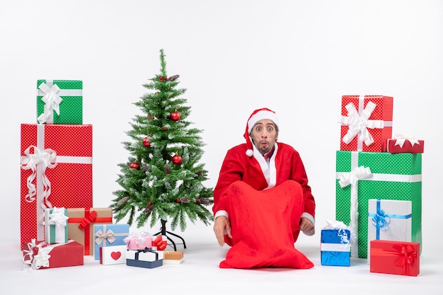 Schockierter junger Mann verkleidet als Weihnachtsmann mit Geschenken und geschmücktem Weihnachtsbaum, der auf dem Boden auf weißem Hintergrund sitzt