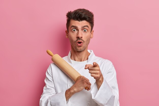 Schockierter männlicher Bäcker hält Nudelholz und zeigt nach vorne, macht sich bereit zum Kochen, arbeitet in der Küche, trägt Uniform