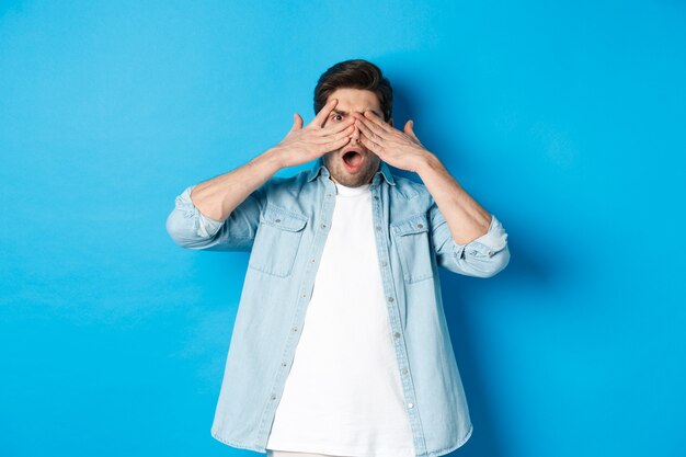 Schockierter Mann, der die Augen bedeckt und durch die Finger späht, starrt auf etwas Peinliches, das vor blauem Hintergrund steht.