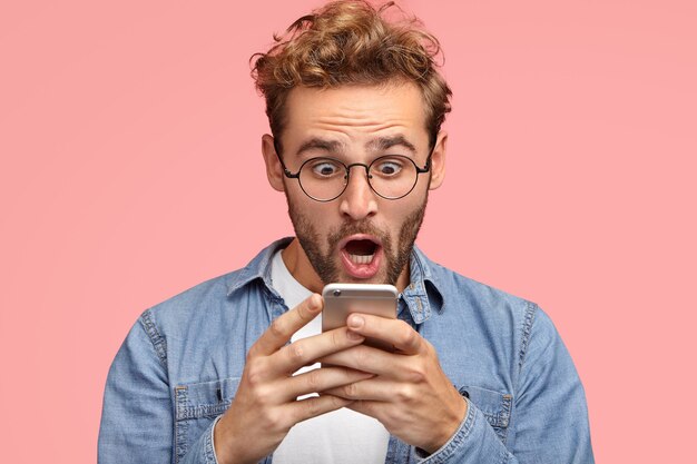 Schockierter unrasierter Mann sieht überraschend aus und mit erschrockenem Ausdruck auf dem Bildschirm des Smartphones, nutzt kostenloses WLAN für Nachrichten in sozialen Netzwerken, liest schreckliche Nachrichten über das Internet. Omg-Konzept