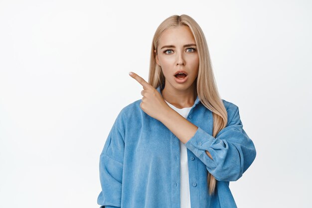 Schockiertes blondes Mädchen, das ehrfürchtig aussieht, mit heruntergelassenem Kiefer, das mit dem Finger auf die obere linke Ecke zeigt und den weißen Hintergrund des Logo-Banners zeigt