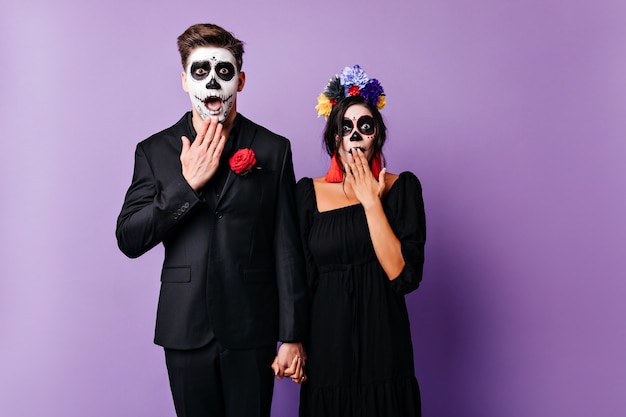 Kostenloses Foto schockiertes europäisches paar mit gruseligem make-up, das hände auf lila hintergrund hält. junge leute in der schwarzen kleidung, die in zombiekleidung in halloween aufwirft.