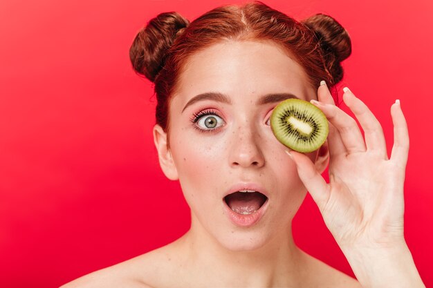 Schockiertes Ingwermädchen, das Kiwi hält. Studioaufnahme der überraschten Frau mit exotischer Frucht lokalisiert auf rotem Hintergrund.