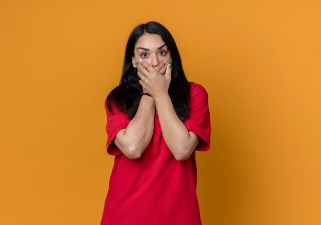 Schockiertes junges brünettes kaukasisches Mädchen, das rotes Hemd trägt, hält Mund mit zwei Händen lokalisiert auf orange Wand