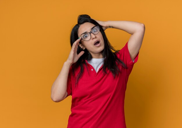Schockiertes junges brünettes kaukasisches Mädchen in der optischen Brille, die rotes Hemd trägt, hält Kopf, der auf orange Wand lokalisiert schaut