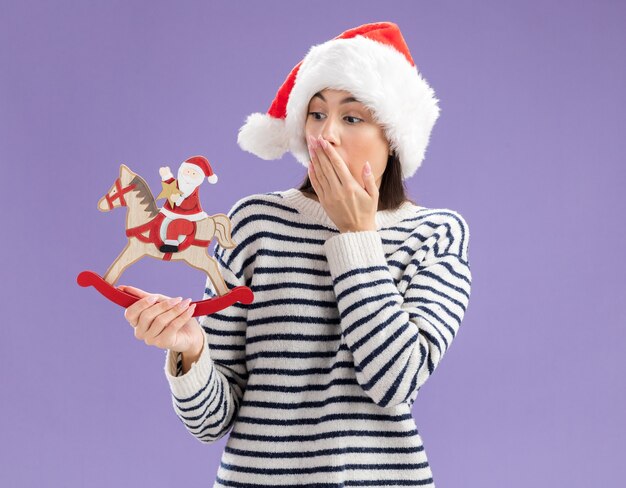 Schockiertes junges kaukasisches Mädchen mit Weihnachtsmütze legt die Hand auf den Mund und sieht den Weihnachtsmann auf Schaukelpferddekoration einzeln auf lila Wand mit Kopierraum an