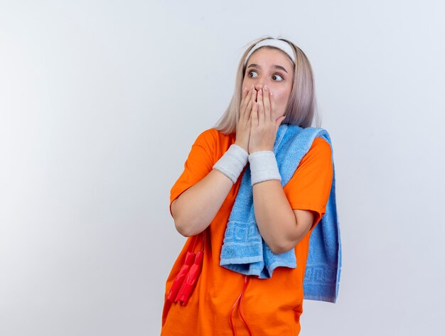 Schockiertes junges kaukasisches sportliches Mädchen mit Zahnspangen und mit springendem Seil um den Hals, das Stirnbandarmbänder hält, die Handtuch auf Schulter halten, legt Hände auf Mund, die Seite auf weißer Wand betrachten