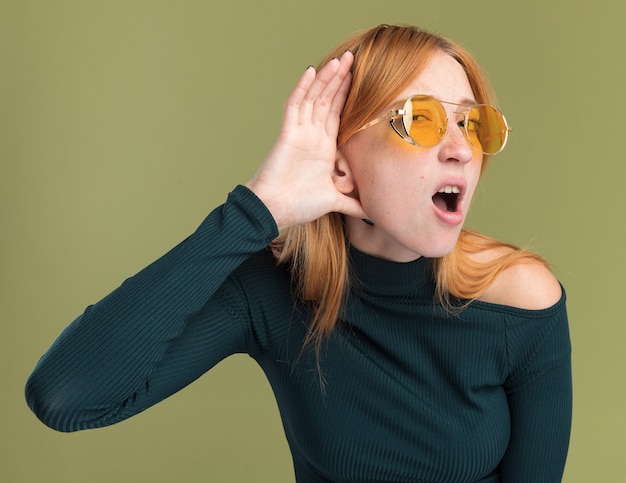 Schockiertes junges rothaariges Ingwermädchen mit Sommersprossen in Sonnenbrille, das die Hand hinter dem Ohr hält, isoliert auf olivgrüner Wand mit Kopierraum
