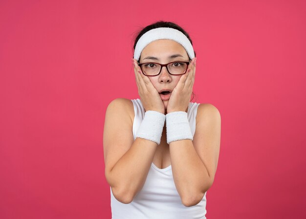 Schockiertes junges sportliches Mädchen in optischer Brille mit Stirnband und Armbändern legt die Hände auf das Gesicht