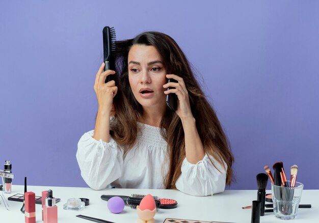 Schockiertes schönes Mädchen sitzt am Tisch mit Make-up-Werkzeugen, die Haare kämmen und am Telefon sprechen, das Seite betrachtet, die auf lila Wand isoliert ist