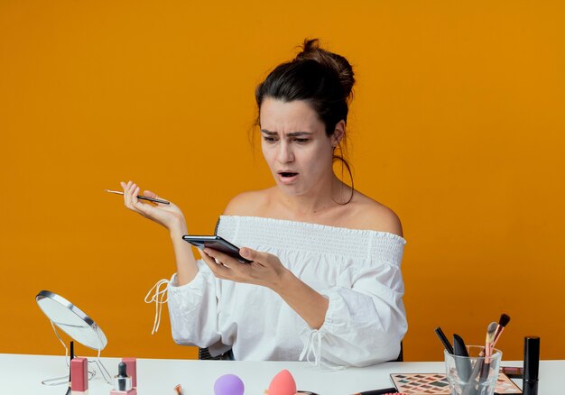 Schockiertes schönes Mädchen sitzt am Tisch mit Make-up-Werkzeugen, die Telefon halten, das Make-up-Pinsel lokalisiert auf orange Wand hält