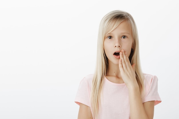 Schockiertes süßes europäisches Mädchen mit blonden Haaren, das Gerüchte oder Klatsch im Klassenzimmer verbreitet und die Handfläche in der Nähe des Mundes hält, während es flüstert, damit niemand anderes ein Geheimnis hört und ernsthaft über der grauen Wand steht