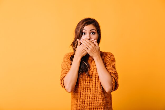 Schockiertes süßes Mädchen, das Mund mit Händen bedeckt. Studioaufnahme der emotionalen kaukasischen Dame lokalisiert auf leuchtendem Orange.