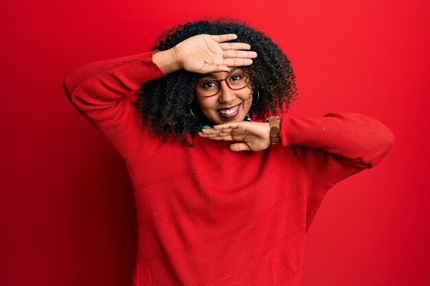 Schöne afrikanisch-amerikanische Frau mit Afro-Haaren, Pullover und Brille, die fröhlich lächelt und Peek a Boo spielt, mit Händen, die Gesicht zeigen. überrascht und begeistert