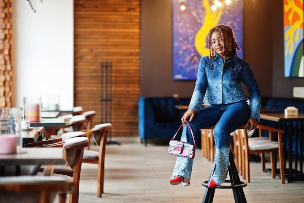 Schöne afrikanisch-amerikanische Frau mit Dreadlocks in blauer, stylischer Jeansjacke im Café Schönes, cooles, modisches, schwarzes junges Mädchen im Innenbereich