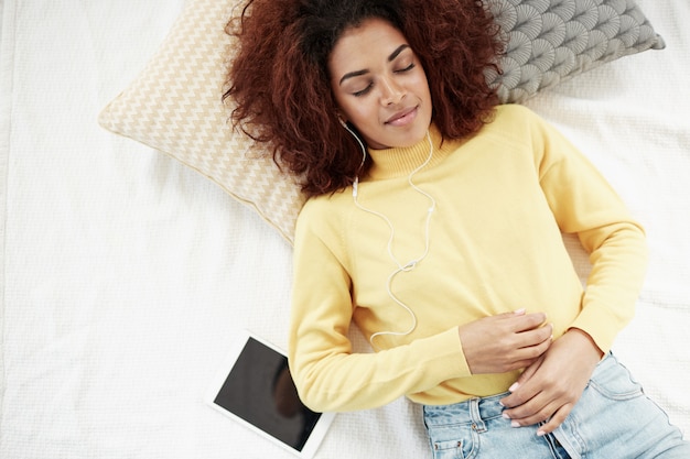 Kostenloses Foto schöne afrikanische frau in den kopfhörern, die musik hören, die auf dem bett liegt, das mit lächelt