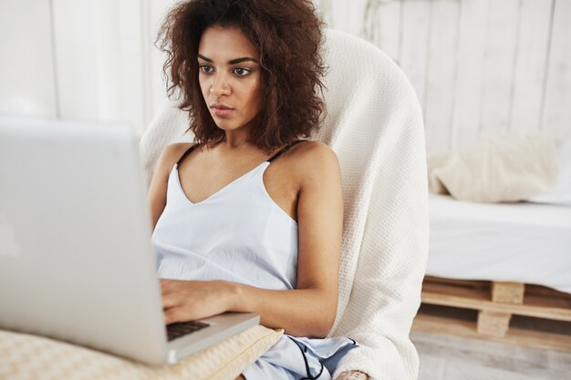 Schöne afrikanische Frau in der Nachtwäsche, die Laptop betrachtet, der im Stuhl zu Hause sitzt.