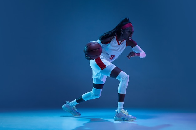 Schöne afroamerikanische Basketballspielerin in Bewegung und Aktion in Neonlicht auf blauem Wandkonzept des professionellen Sporthobby des gesunden Lebensstils