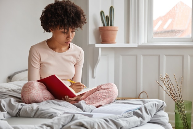 Schöne afroamerikanische Frau sitzt auf dem Bett, kreuzt die Beine, macht einige Notizen im Tagebuch hält Stift in Händen