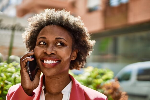 Schöne afroamerikanische Geschäftsfrau mit Afro-Haaren, die draußen in der Stadt glücklich und selbstbewusst lächelt und ein Gespräch am Telefon führt