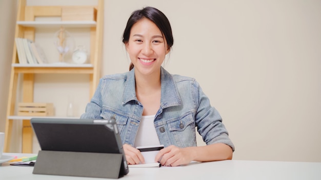 Schöne asiatin, die die tablette kaufen online kaufen mit kreditkarte beim zufälligen sitzen der abnutzung auf schreibtisch im wohnzimmer zu hause verwendet.