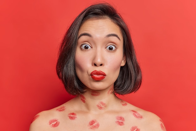 Schöne asiatische Dame mit roten Lippen posiert mit nackten Schultern gegen leuchtend rote Wand hat überraschte Ausdruckskussspuren auf Körperposen im Innenbereich