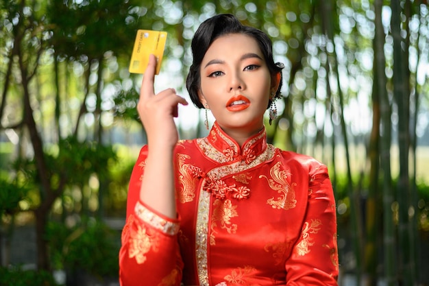 Schöne asiatische Frau des Porträts in einer chinesischen cheongsam-Pose mit Kreditkarte auf Bambuswald