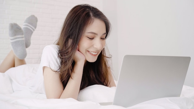 Schöne asiatische Frau, die Computer oder Laptop beim Lügen auf dem Bett in ihrem Schlafzimmer spielt.