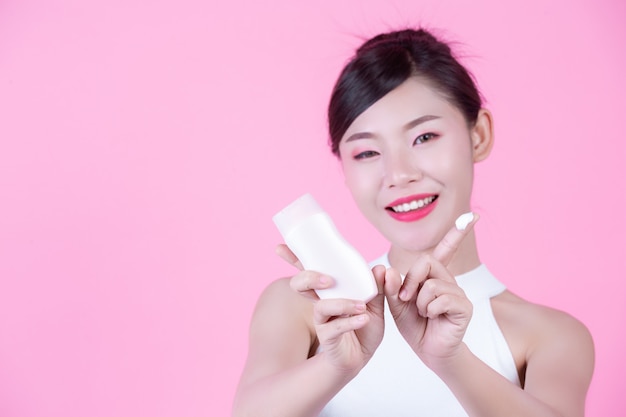 Schöne asiatische Frau, die eine Flasche des Produktes auf einem rosafarbenen Hintergrund anhält.