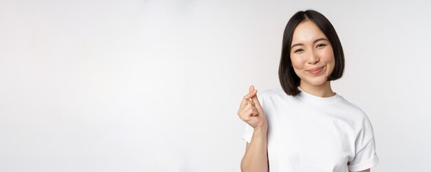 Schöne asiatische Frau, die lächelt und Fingerherzen zeigt, die ein T-Shirt tragen, das gegen weiße Ba steht