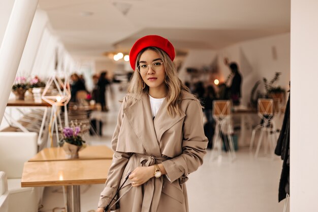 Schöne asiatische Frau im trendigen Trenchcoat und in der hellen Baskenmütze hält Tasche und schaut nach vorne gegen gemütliches Zimmer