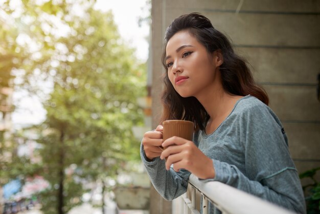 Schöne asiatische Frau mit der Tasse Tee schauend aus ihrem Balkon heraus