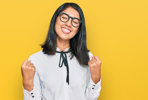 Schöne asiatische junge Frau trägt Business-Hemd und Brille, sehr glücklich und aufgeregt, während sie mit erhobenen Armen lächelnd und schreiend nach dem Erfolgsfeierkonzept eine Siegergeste macht